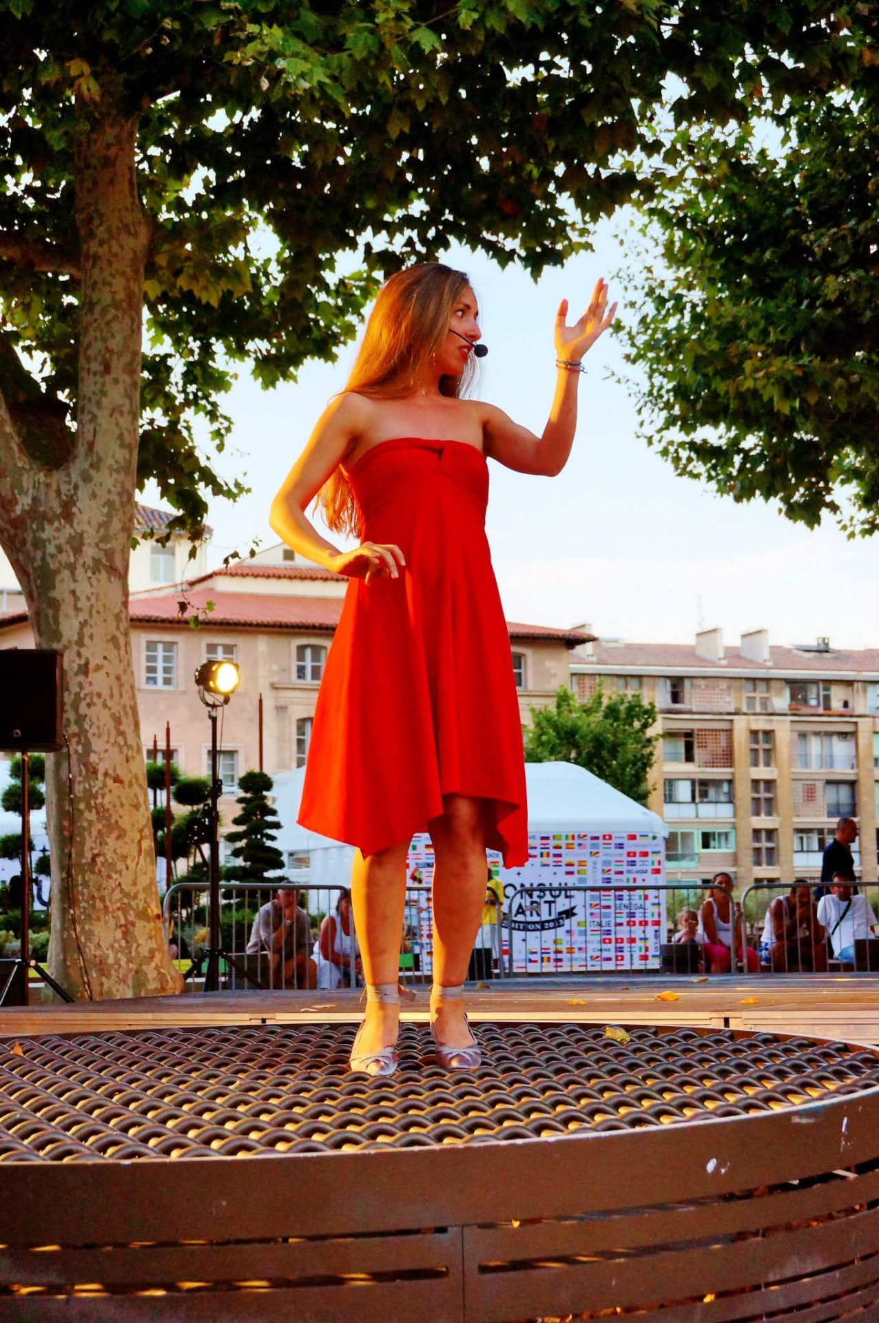 Elisabeth Theodorou performing in a red dress on an open-air stage beneath a plane tree